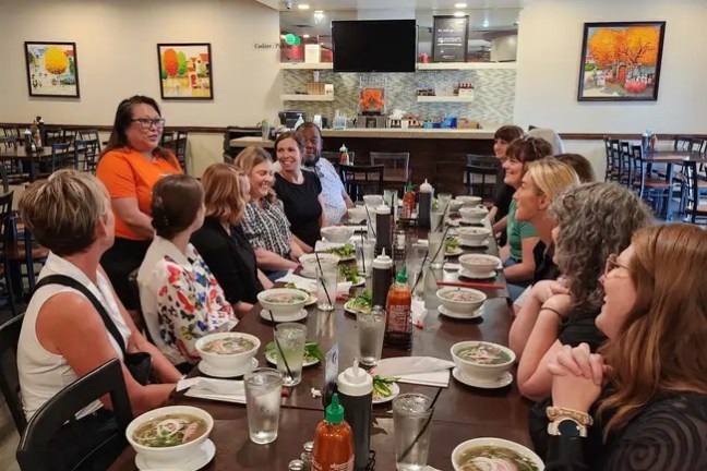 Group of people seated at a long table in a restaurant, bowls of soup and condiments visible.