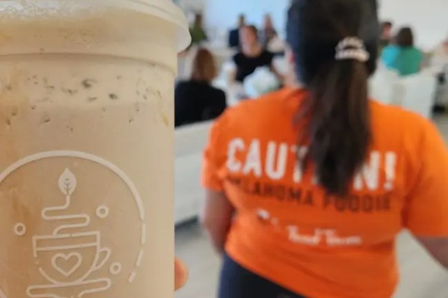 Close-up of bubble tea cup, person in orange 'Caution! Oklahoma Foodie' shirt in blurred background.
