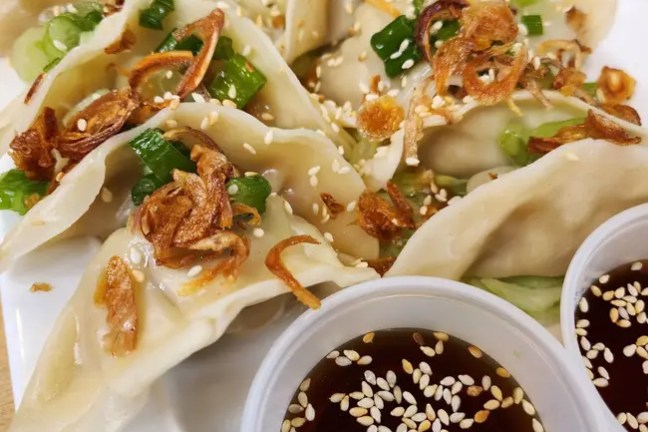 Plate of dumplings topped with scallions and fried onions, with dipping sauce cups.
