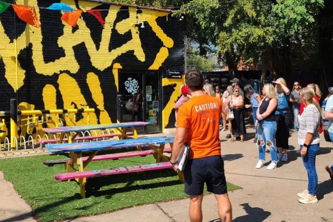 People gathered outside building with bold mural and picnic tables.