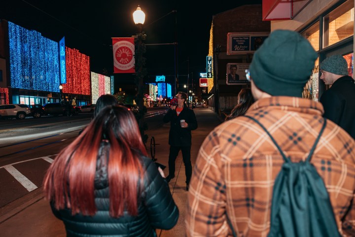 People walking on a lit city street at night, colorful building lights in the background.