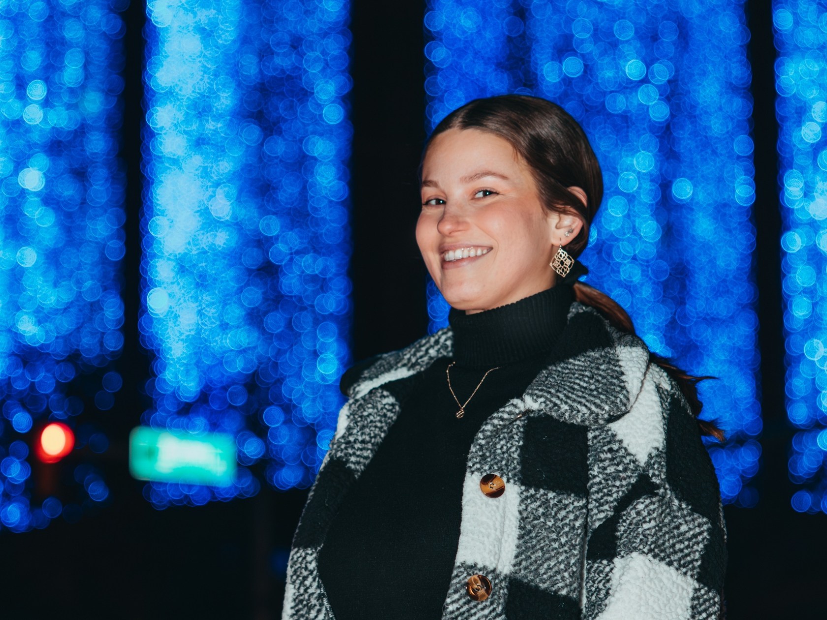 Woman in checkered jacket smiles at night with blue lights in background.