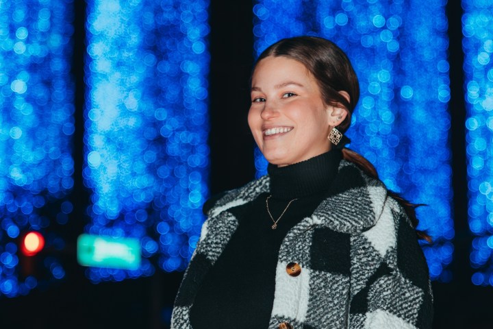 Woman in checkered jacket smiles at night with blue lights in background.