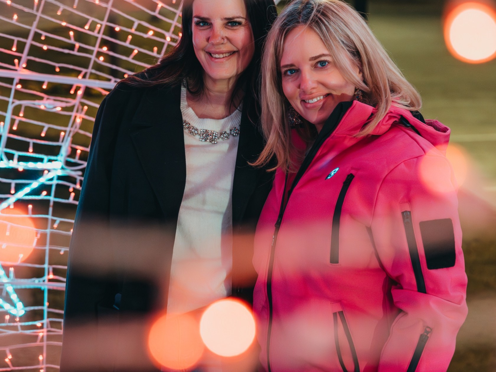 Two women smiling under arch with festive lights and blurred red bokeh effect.
