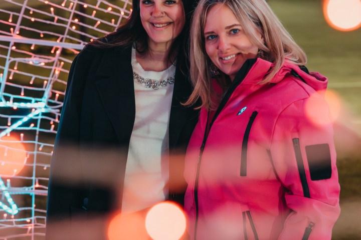 Two women smiling under arch with festive lights and blurred red bokeh effect.
