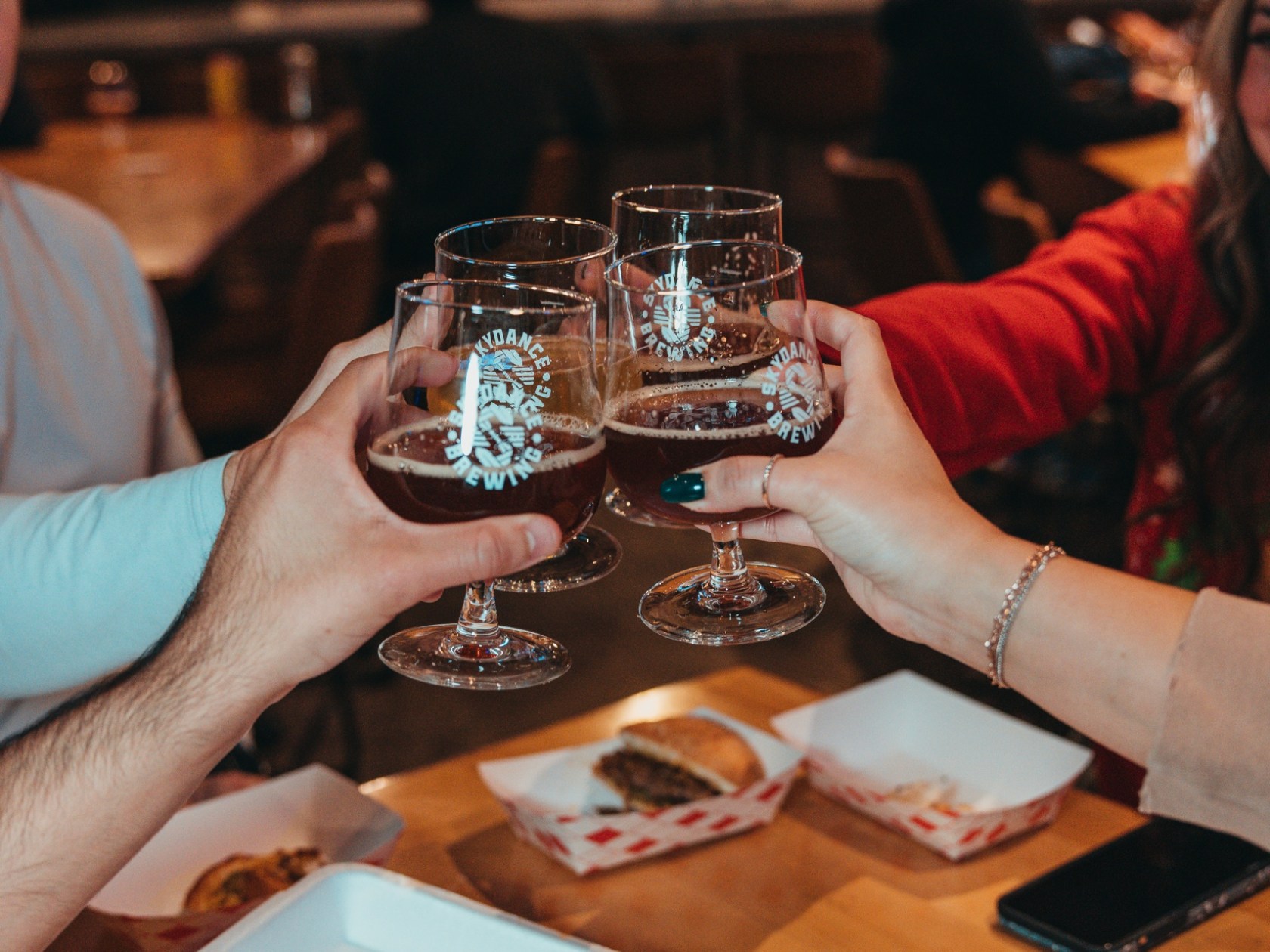 Four hands toasting with beer glasses over food in a restaurant.