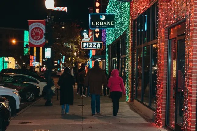 People walking on a city sidewalk at night with colorful lights and signs.